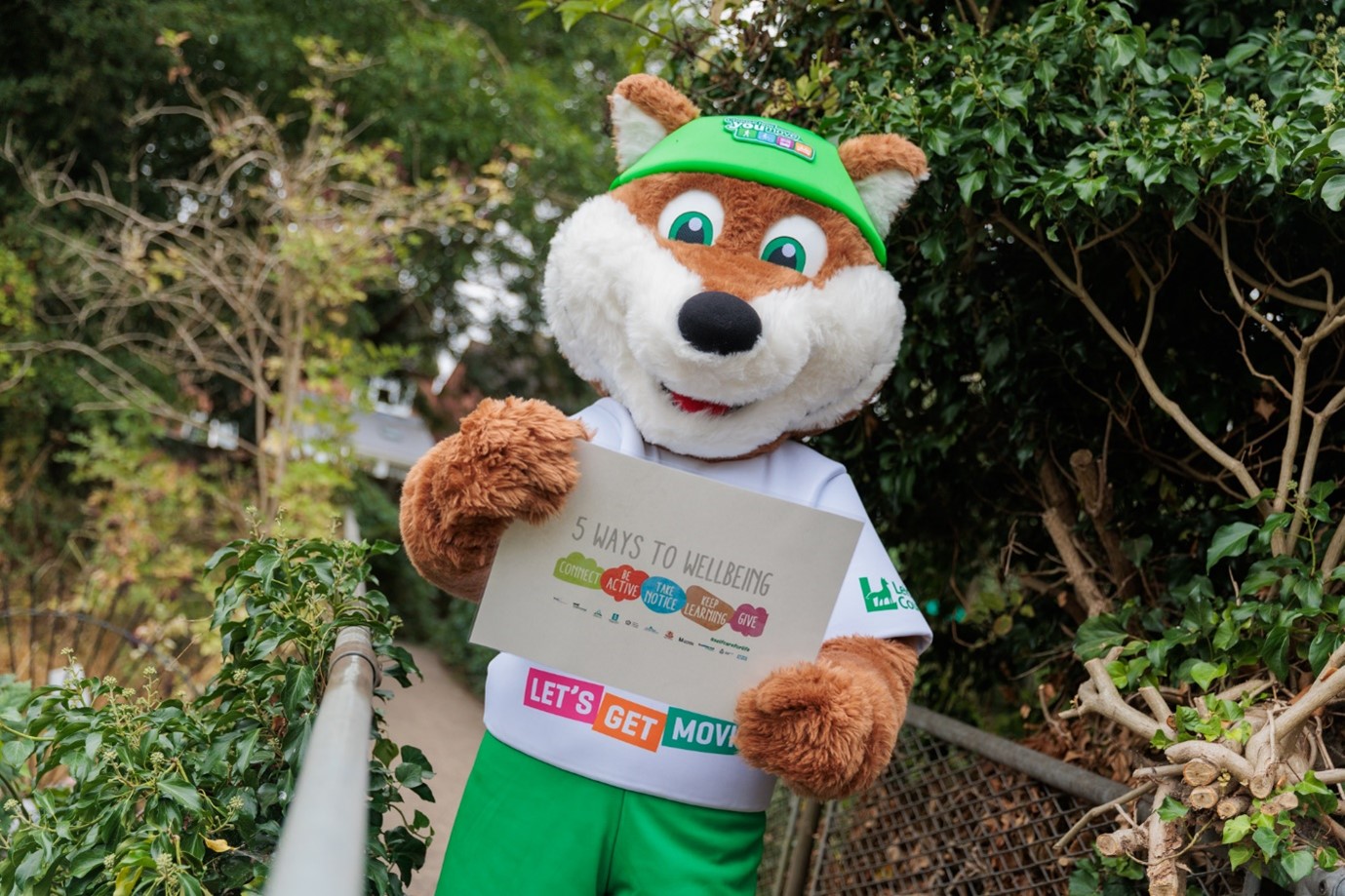 Theo the Fox, the mascot for Choose How You Move, stands in front of a public footpath surrounded by trees and bushes, and holds up a laminated piece of paper displaying the '5 Ways to Wellbeing'