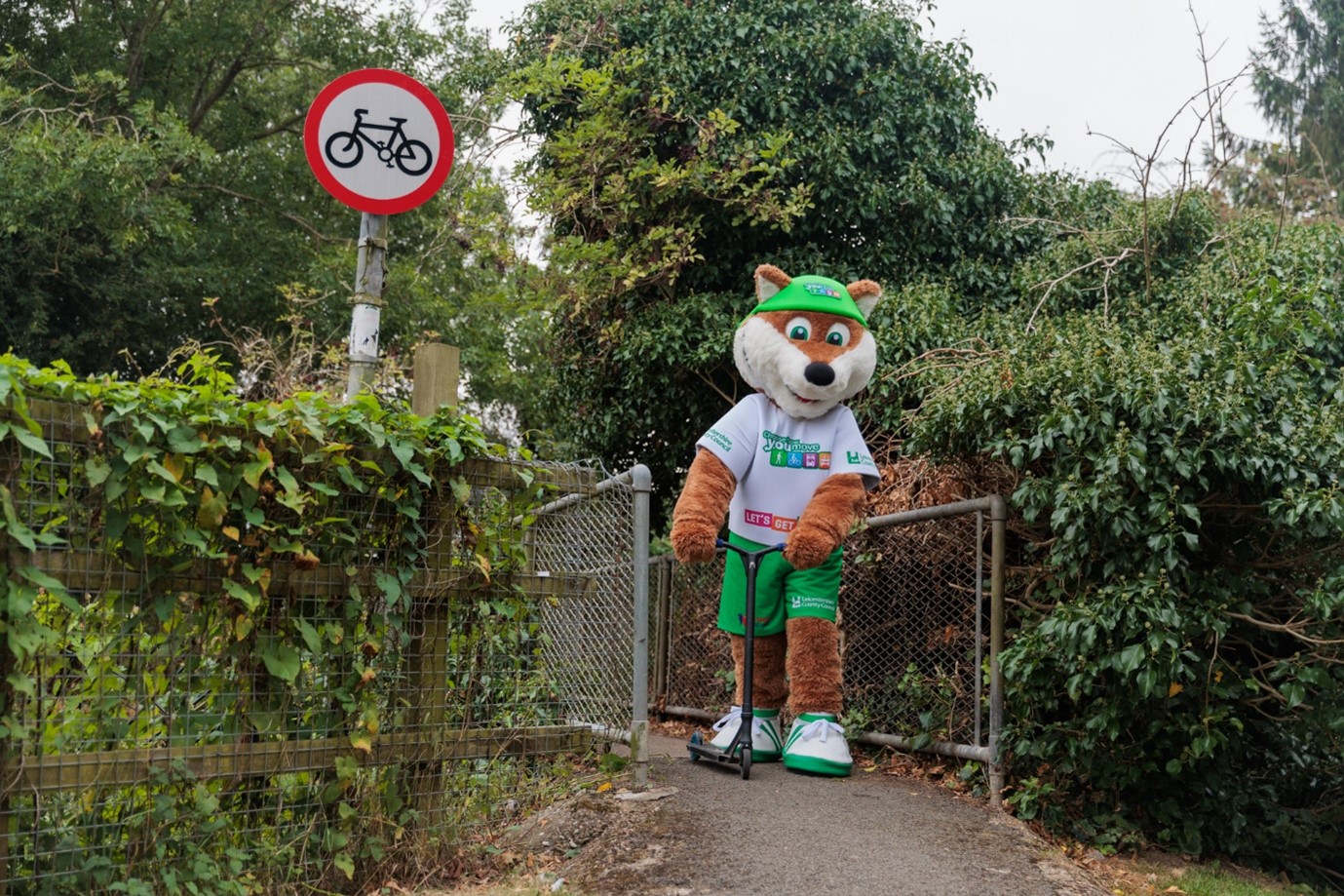 Theo the Fox, the Choose How You Move mascot, stands with a scooter in front of a public footpath as part of a back to school active travel photoshoot. To his left is a sign indicating no bicycles.