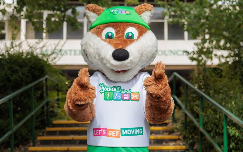 Theo the Fox, a Choose How You Move mascot, stands in front of County Hall and makes finger guns